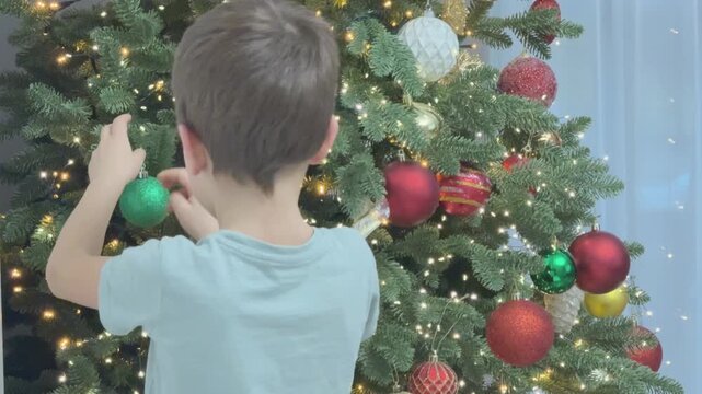 Boy hanging green ornament on Christmas tree with focus on decoration process. Warm festive home atmosphere.
