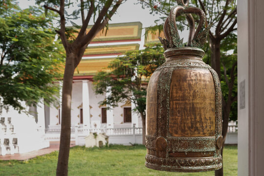A traditional Thai-style bell hangs with a tree and a Thai temple ordination hall in the background.