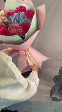 woman arranges pink paper wrapped bouquet on fabric covered tabletop in small indoor studio corner, hands tying satin ribbon and carefully adjusting flowers by hand