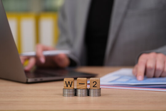 At a modern office desk, a person prepares for tax season with W-2 forms and neatly stacked coins. A laptop and financial papers are in focus, symbolizing organization.