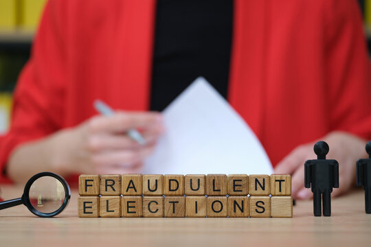 A person dressed in red sits at a table with wooden blocks spelling out FRAUDULENT ELECTIONS. Nearby are mini figures and a magnifying glass, hinting at analysis of election integrity.