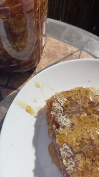 close-up motion shot of honeycomb piece and nearby jar with amber honey visibly pooling, dripping, and oozing along jar rim and plate edge, strong overhead, with