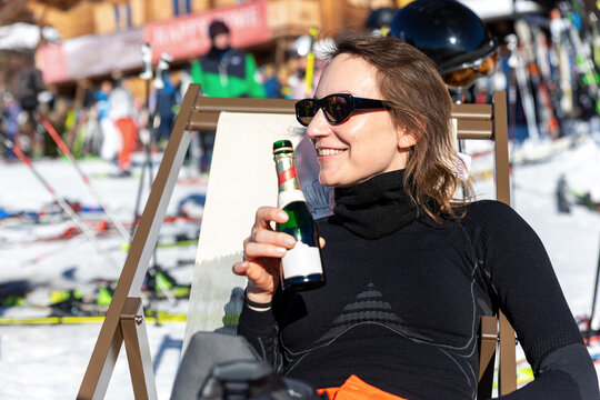 Smiling woman in sunglasses relaxes on deck chair at snowy ski resort, holding beer bottle beside slopes and equipment. Sunny winter scene suggests apres ski leisure, vacation, sport, and celebration