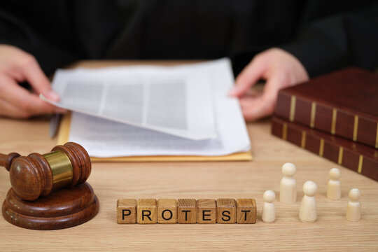 A judge examines legal documents while a gavel rests nearby. Wooden letters spell out PROTEST, symbolizing the theme of dissent in a legal context, focusing on civic engagement.