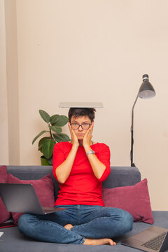A person sits cross-legged on a sofa at home with a laptop on one side and a laptop balanced on their head