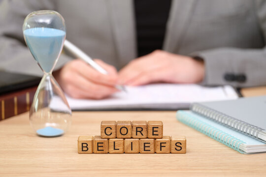 In a serene workspace, a person pens thoughts while an hourglass marks time. Stacked wooden blocks spell out core beliefs, encouraging introspection and growth.