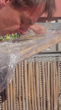 close-up motion of forearm and hand peeling clear plastic wrap from large round baked cake resting on bamboo tray on balcony, steady grip supports tray underside