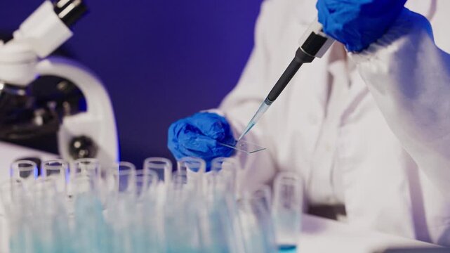 Scientist in protective gear using pipette to transfer blue liquid into test tubes in modern laboratory