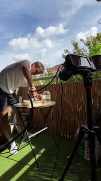 man leaning over small balcony table manipulating honeycomb on wrapped board while camera on tripod records close-up, hands press and slice honeycomb next to glass