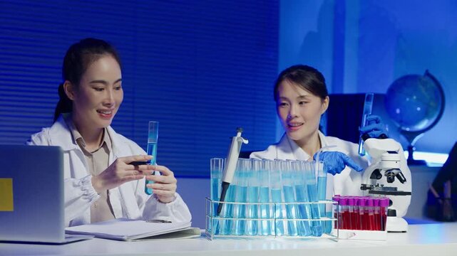 Two Asian Female Scientists Working Together in Modern Blue Laboratory with Test Tubes and Microscope