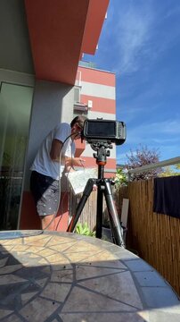 man in white t-shirt and dark shorts adjusts monitor mounted on tripod while holding white reflector panel and cables on urban apartment balcony, low-angle, with