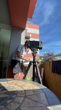 man on apartment balcony grips framed glass panel with pliers while camera monitor mounted on tripod occupies foreground, scene shows mosaic table in lower, with