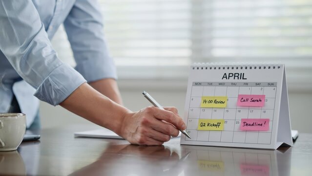 A person in a blue shirt writes on a desk calendar for april with colorful sticky notes for 14:00 review, call sarah, and project deadline