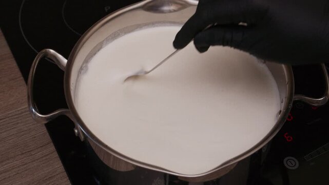High angle shot of chef hands stirring hot milk in stainless steel pot on induction stove. Cooking process for homemade cheese, sauce, or warm beverage.