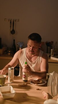 Vertical video of middle aged Caucasian man opening beer bottle and drinking alcohol at table. Footage fits mental health awareness campaigns and addiction recovery content
