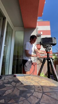 man stands on apartment balcony handling and aligning wooden slats while adjusting tripod-mounted camera, suggesting setup for diy or instructional recording, with