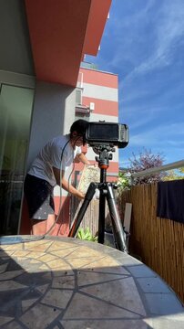 man on residential balcony leans over tripod-mounted monitor while holding translucent honeycomb or glass object near device, low-angle viewpoint from patterned