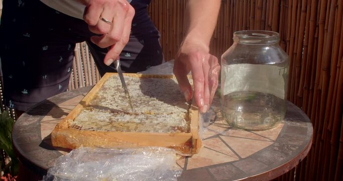 motion shows hands slicing capped honeycomb from wooden board, steadying pieces while lifting cut portion toward empty glass jar on mosaic table in bright sunlight