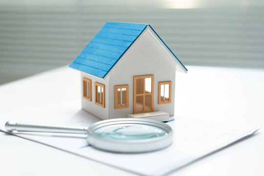 Miniature house with magnifying glass and documents represents property inspection, home valuation, real estate research and careful assessment before buying or investing in residential property.