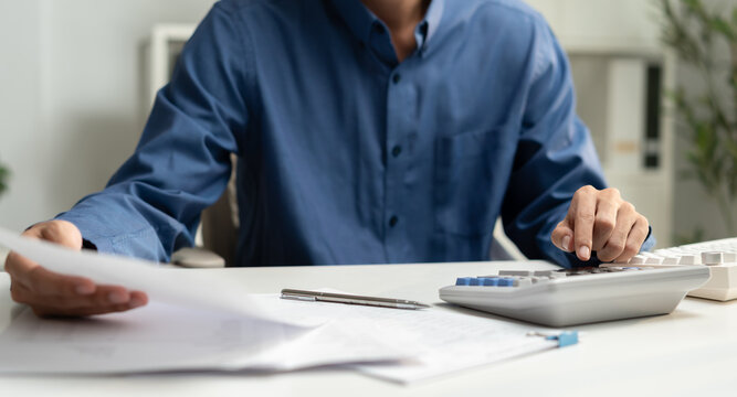 A professional male businessman in a blue shirt calculates financial data using a calculator while reading paperwork and accounting documents at his modern office desk.
