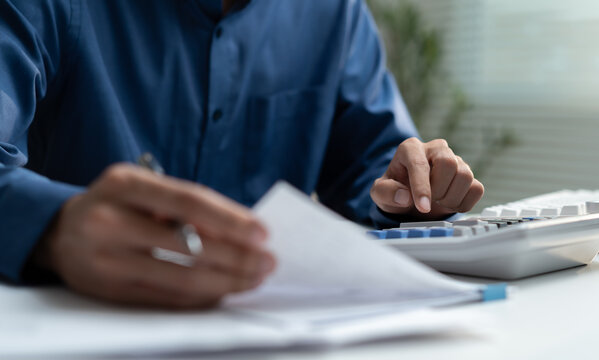 A male accountant holds a pen to review paperwork and financial documents while using a calculator on the desk. The businessman audits tax reports for company budget planning.
