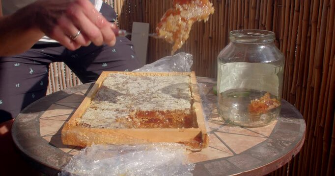 close-up hands cut and lift honeycomb, then place wax comb into glass jar on outdoor patio table, bathed in warm sunlight and golden honey, sticky amber texture