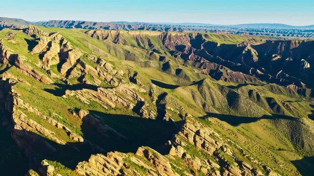 Aerial view of a vast canyon with layered rock cliffs, green ridges, deep shadows, and bright sunlight beneath a clear blue sky.