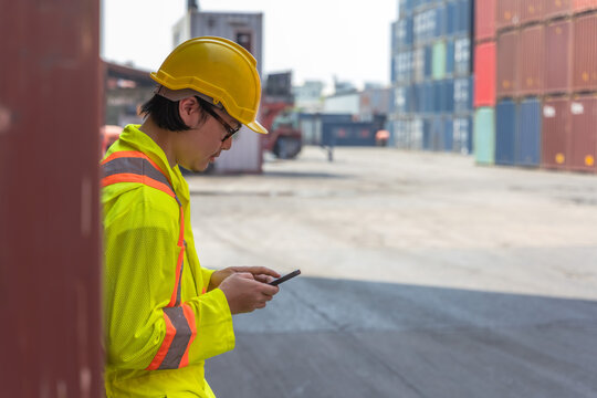 Asian male port terminal worker relaxing and looking at phone during break after finished work inspecting shipping containers stored in a crowded container yard for international export