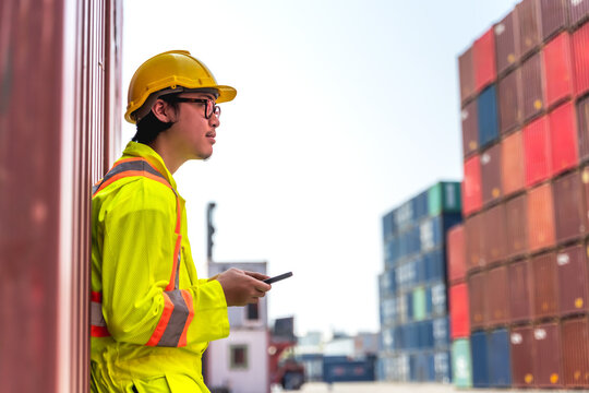 Asian male port terminal worker relaxing and looking at phone during break after finished work inspecting shipping containers stored in a crowded container yard for international export