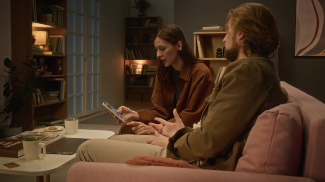 Young Caucasian couple using smartphone and discussing digital content together indoors. Clip fits commercial advertising for mobile applications, fintech platforms, and ecommerce services