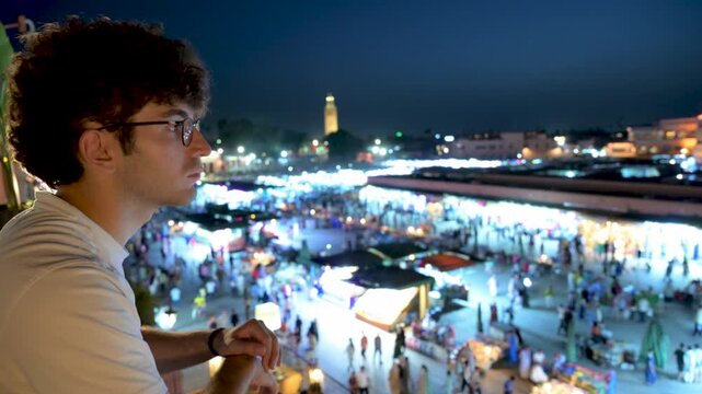 Marrakech, morocco. August 9, 2026. Thoughtful young man with glasses admiring the panoramic night view of the bustling jemaa el fnaa square in marrakech, morocco from a rooftop