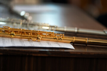 Golden flute rests beside silver one on wooden surface. Sheet music lies beneath, suggesting rehearsal or performance. Soft focus blurs background, emphasizing instrument details © Natalia