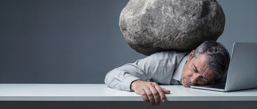 Overworked businessman slumped beside a laptop while a massive boulder weighs on his shoulders, visual metaphor for extreme workload, stress, burnout and professional pressure, copy space.