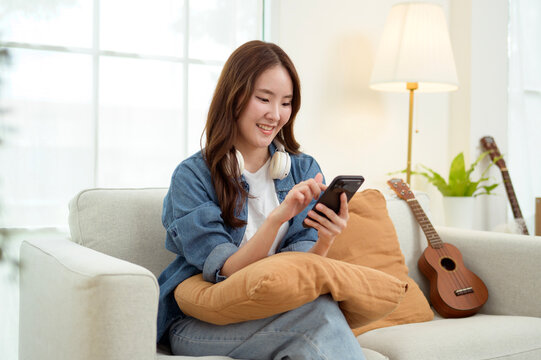 Asian Woman Using a Smartphone While Sitting on a Couch With a Pillow in a Bright Room