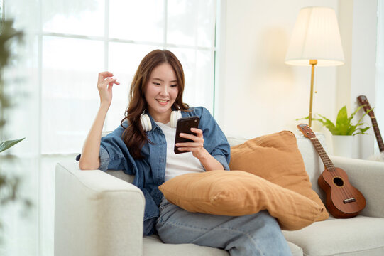 Asian Woman Using a Smartphone While Sitting on a Couch With a Pillow in a Bright Room