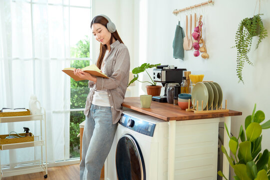 Young Woman Reads Book While Standing Next to Washing Machine in Home Kitchen