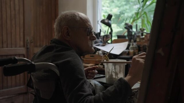 Senior artist on a wheelchair paints an oil painting on canvas in a studio