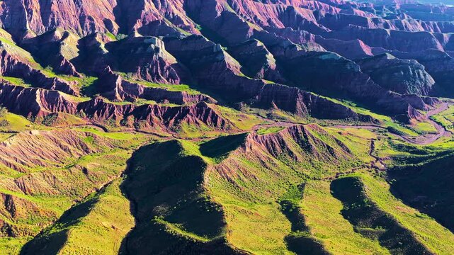 Aerial view of rugged eroded mountain ridges with layered rock formations, green vegetation, deep ravines, and strong sunlight across the terrain.