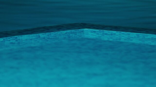 Illuminated swimming pool with a blue tile border and a blue water surface, night shot. The water is calm and still
