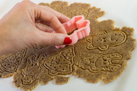 Close-up of a woman's hand with red fingernails using a pink cookie cutter to outline festive shapes on stamped gingerbread dough.