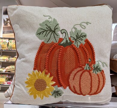 Tiempo de oto&ntilde;o. Coj&iacute;n con dibujo de calabazas y flor de girasol, con ribete marr&oacute;n, en estante de tienda de decoraci&oacute;n 