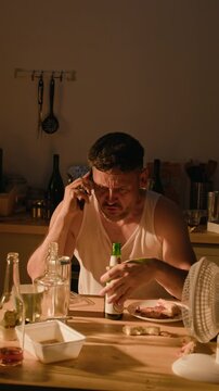 Vertical shot of middle aged Caucasian man arguing on smartphone and drinking beer at messy table. Footage suitable for substance abuse awareness, addiction rehabilitation and mental health campaigns