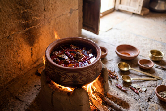 Traditional Rajasthani laal maas mutton curry simmering in an earthen clay pot over an open fire, with red chilies and whole spices in a rustic village kitchen in Rajasthan, India