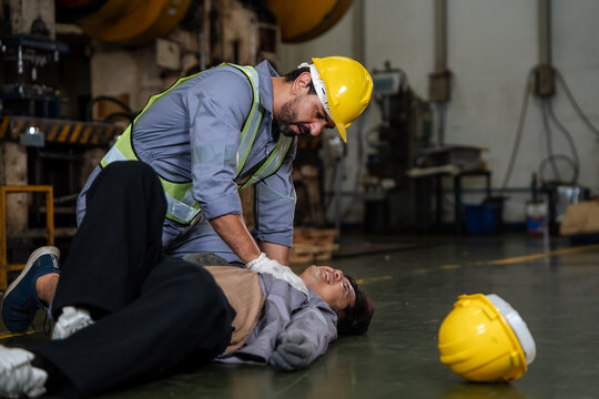 Worker performs CPR on a person lying on factory floor. He wears yellow hard hat and safety vest. Industrial machinery with large gears is visible. Spare hard hat rests on floor