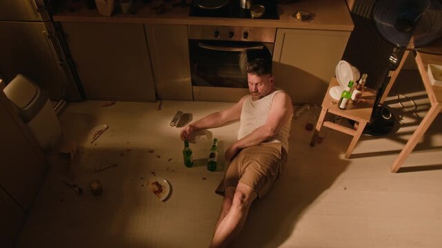 Caucasian young adult man lying intoxicated on kitchen floor, filmed from high angle while drinking beer. Ideal for addiction recovery and mental health campaigns