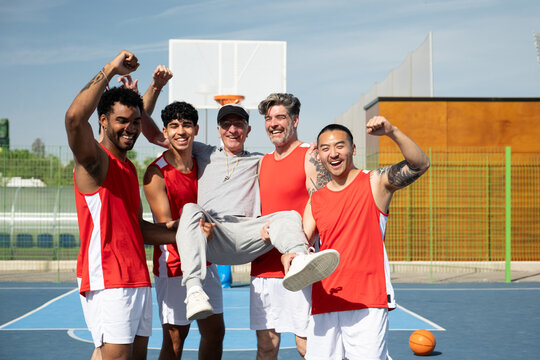 Diverse basketball players cheering and smiling, carrying their coach after a winning game