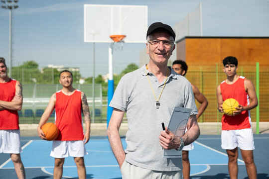 Coach holding clipboard and whistle with team on an outdoor basketball court
