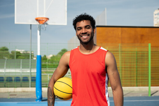 Young man smiling at camera holding a basketball on an outdoor court