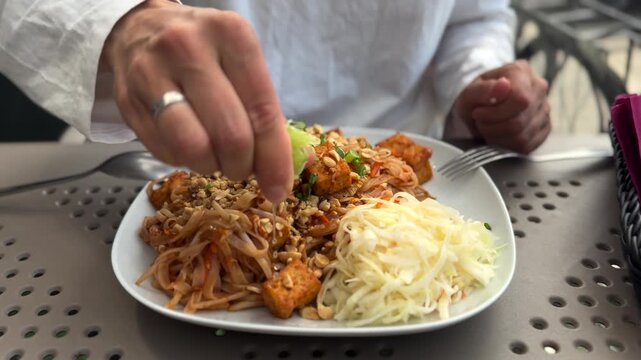 Hand squeezes lime over plated noodles and tofu beside shredded cabbage, adding fresh citrus to richly garnished meal on outdoor table. Finishing touch emphasizes flavor and ready to eat presentation.