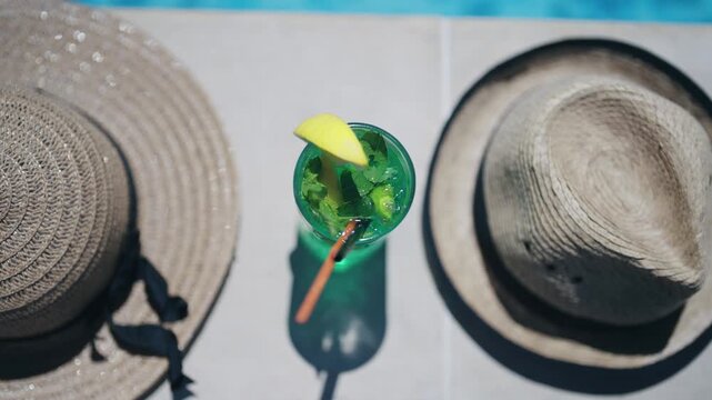 A green coctail drink with a straw in it is sitting on a ledge next to a pool. The drink is in a tall glass and has a lime wedge in it. Concept of relaxation and leisure